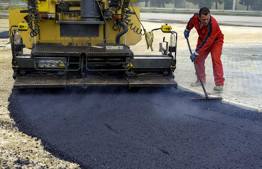 Road construction worker smoothing plant mix asphalt to form the surface of a newly constructed road as part of a public infrastructure development project. 